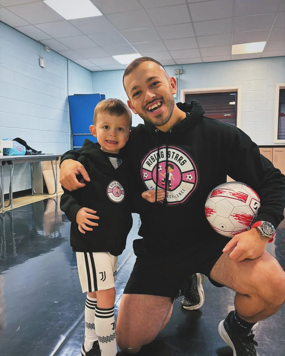 A smiling man and a young boy, both wearing matching black hoodies with a pink and white logo, stand together in an indoor gym. The man is kneeling on one knee, holding a soccer ball, and the boy is hugging the man. The gym has a blue wall, a table with bags and items, and a reflective black floor.