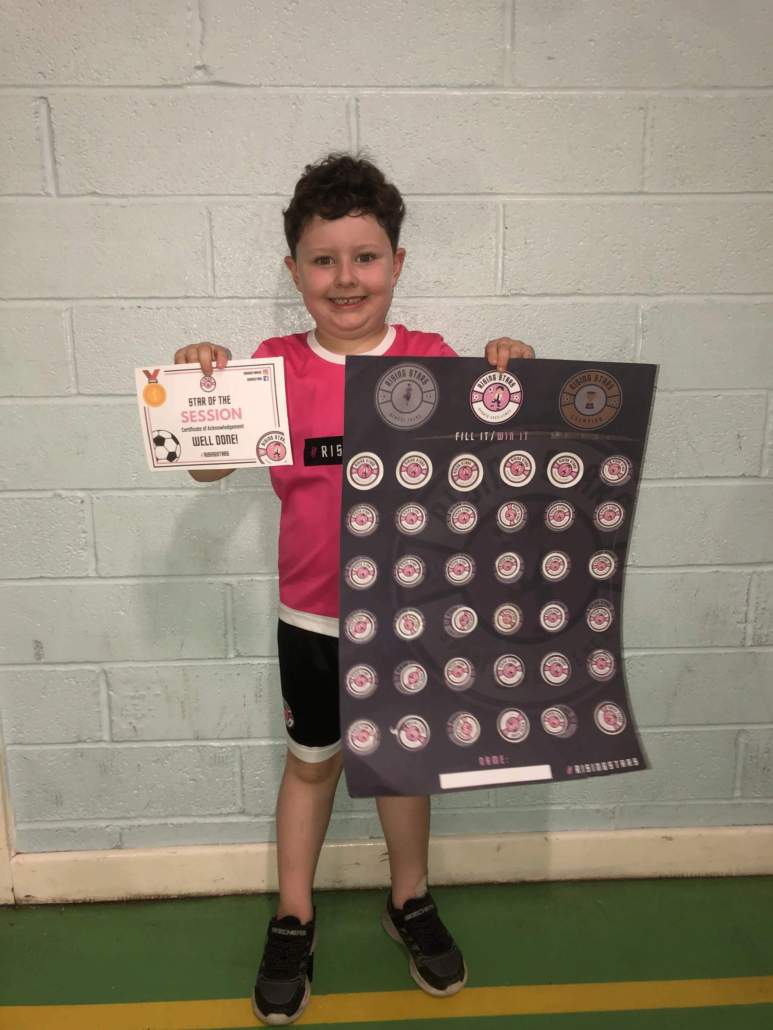 A young boy standing in front of a white brick wall, holding a certificate and a poster, smiling at the camera. The certificate reads 'Star of the Session' and the poster features multiple pink and black icons with a theme related to a sports team.