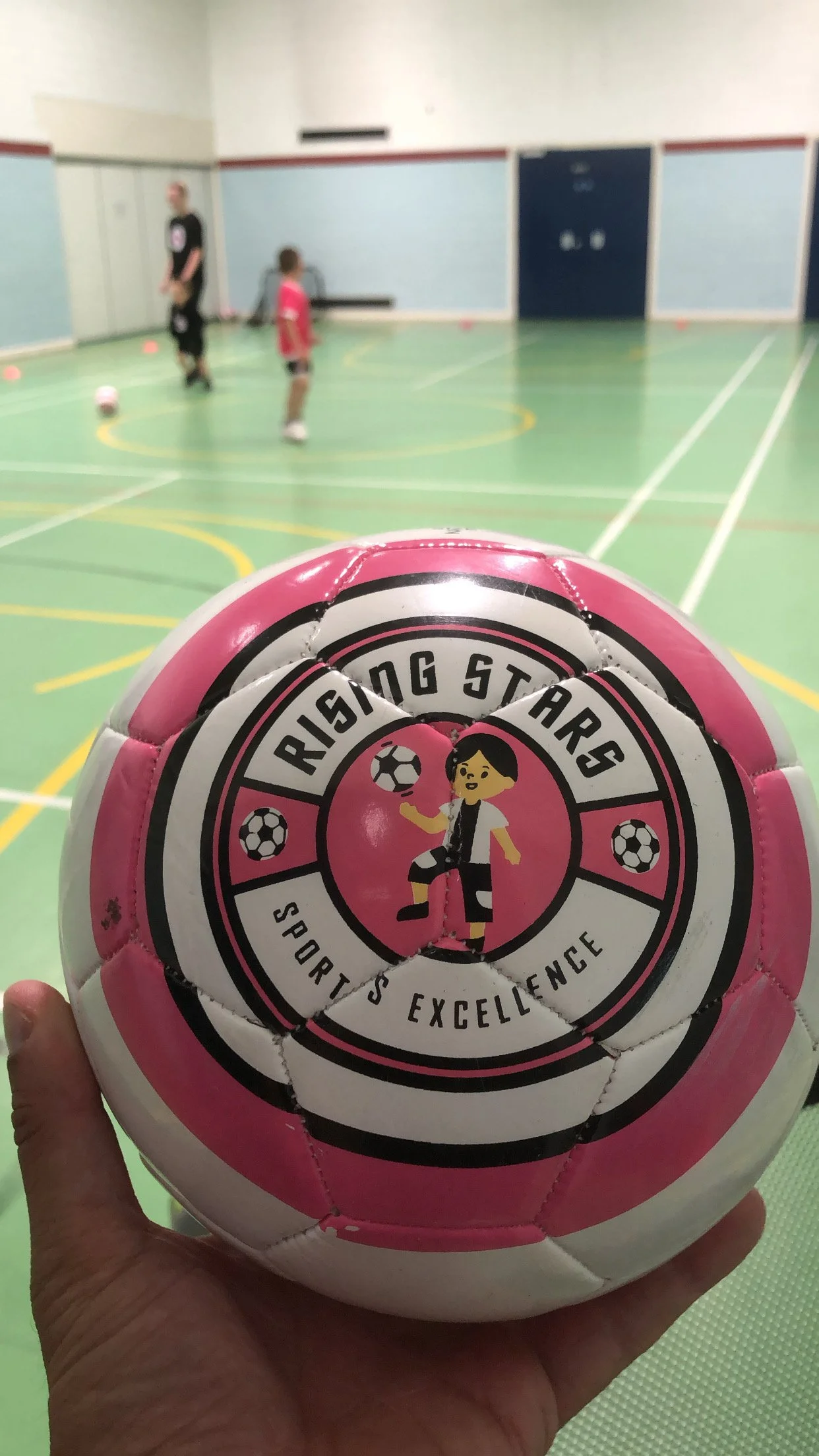 Pink and white soccer ball with a logo reading 'Rising Stars Sports Excellence' held in a gymnasium with children practicing soccer in the background.