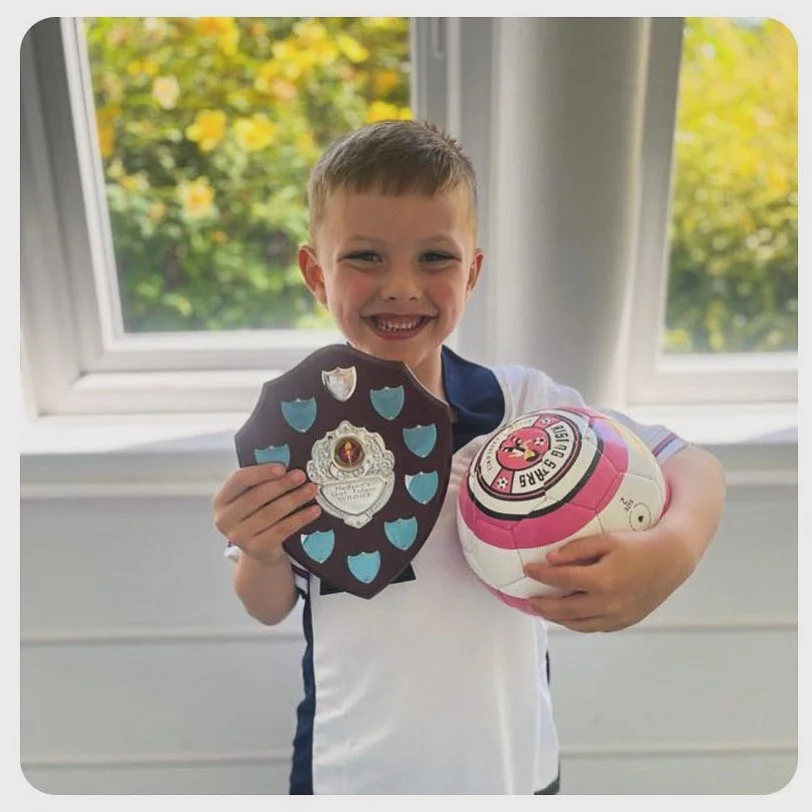 A young boy with a big smile holding a badge and a pink and white sports ball, standing in front of a window with greenery outside.