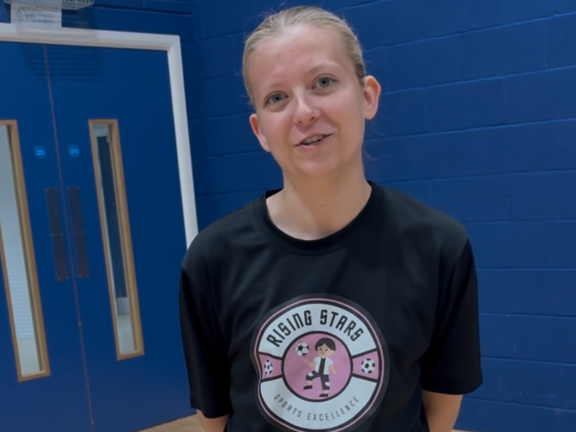 A young girl with blonde hair tied back, smiling, standing in front of a blue gymnasium wall, wearing a black T-shirt with a sports logo for Rising Stars Sports Excellence.