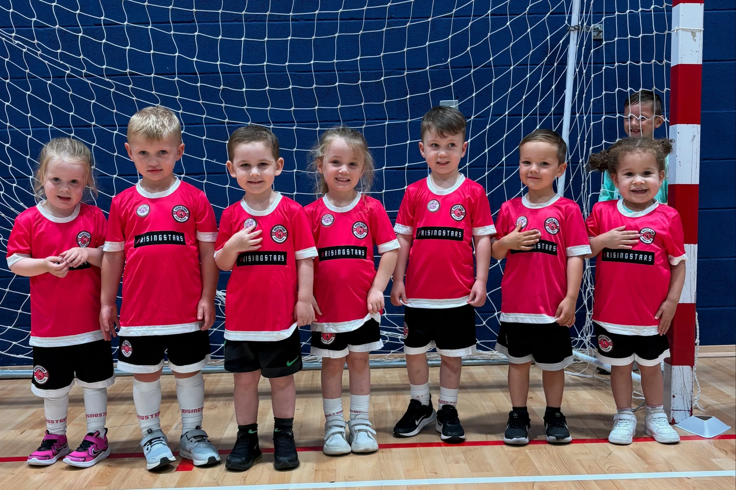 Children in red soccer jerseys and black shorts standing in front of a goal net in an indoor gym.