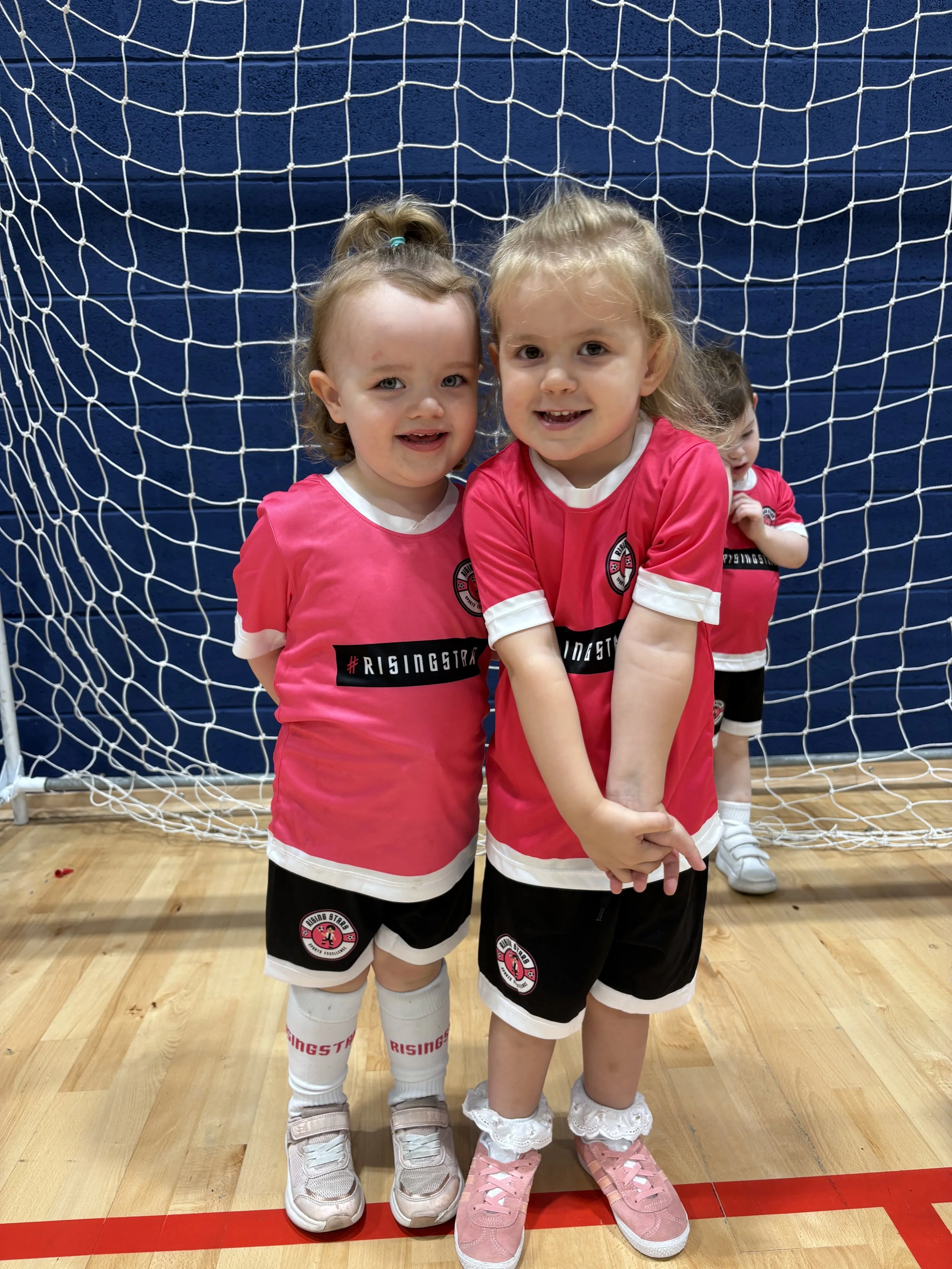 Three young girls in pink and black soccer uniforms standing in front of a soccer goal on an indoor court.