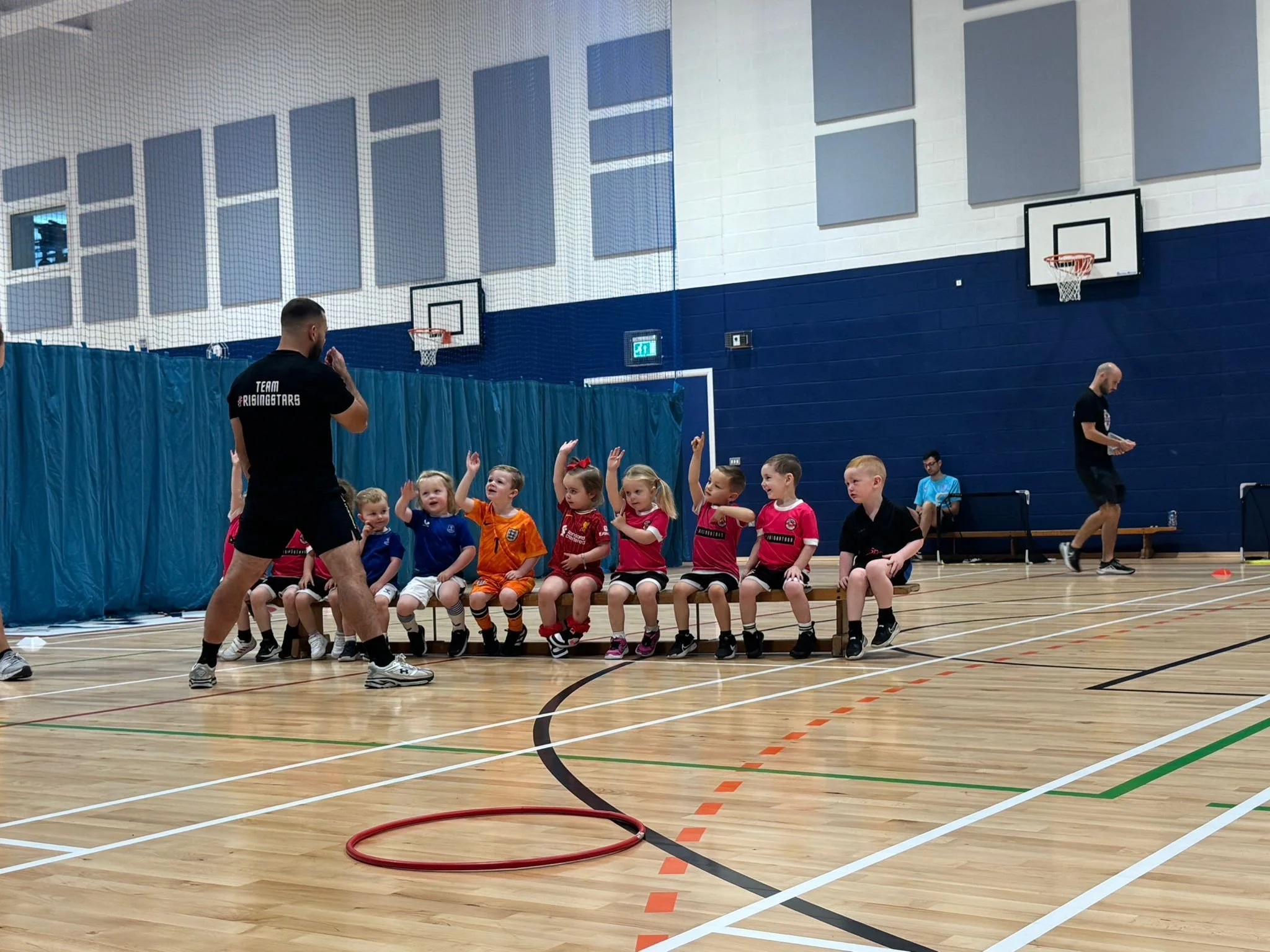 Children sitting on a bench in a gymnasium during a sports training session, with a coach standing in front of them and another person walking nearby.