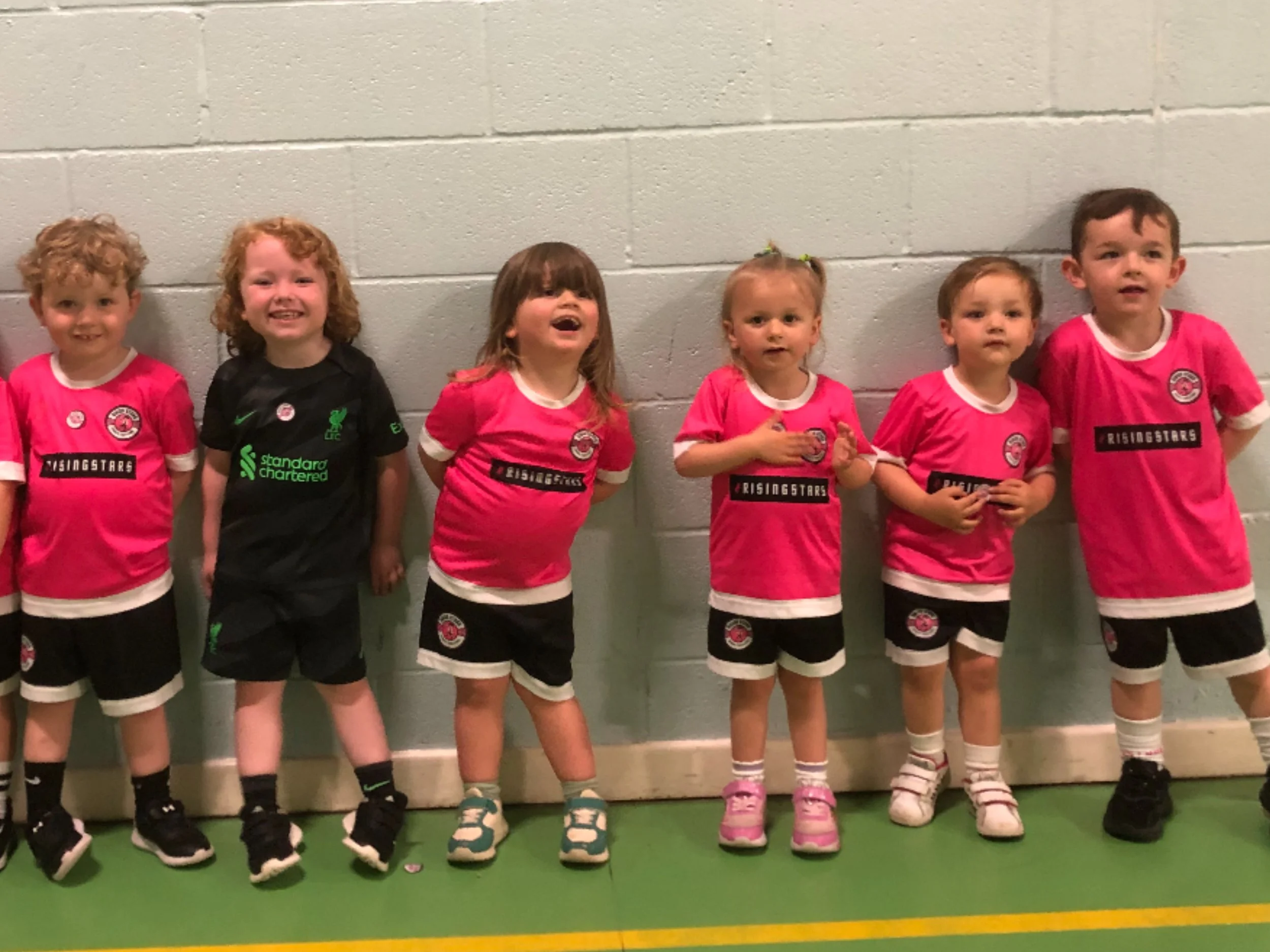Six young children standing in a row against a gray wall, dressed in pink soccer uniforms with one child in black, smiling and looking at the camera.