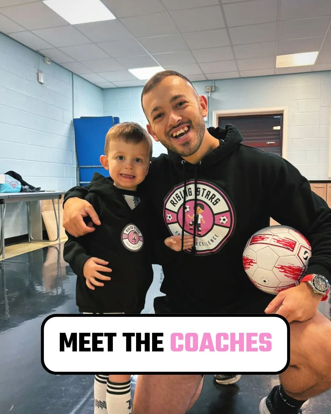 A man and a young boy are smiling and hugging, both wearing black sports hoodies with a pink and white logo that says 'Rising Stars Soccer Excellence.' The man is holding a soccer ball. The setting appears to be an indoor sports facility or gym.