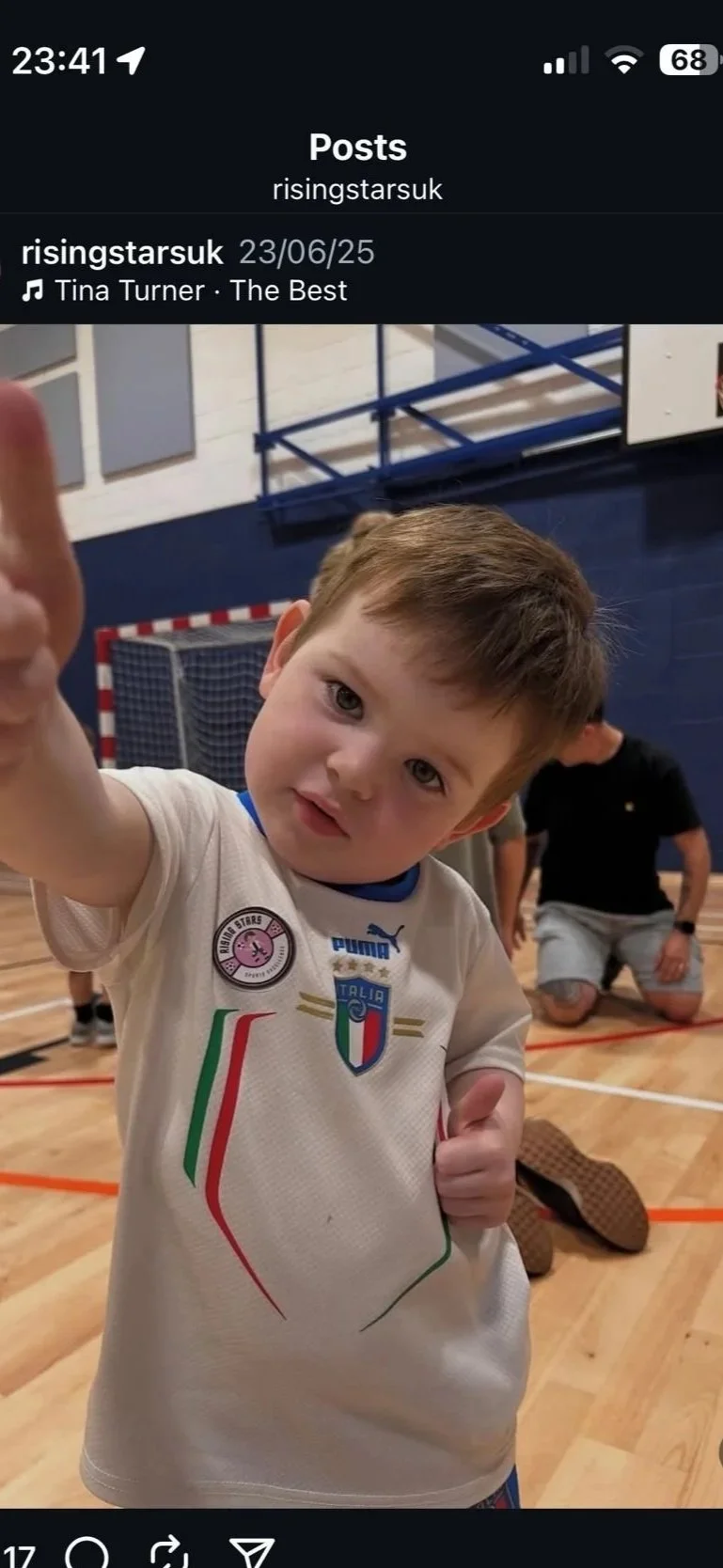 Young boy in a sports uniform pointing at the camera in an indoor gymnasium with a goalpost in the background.