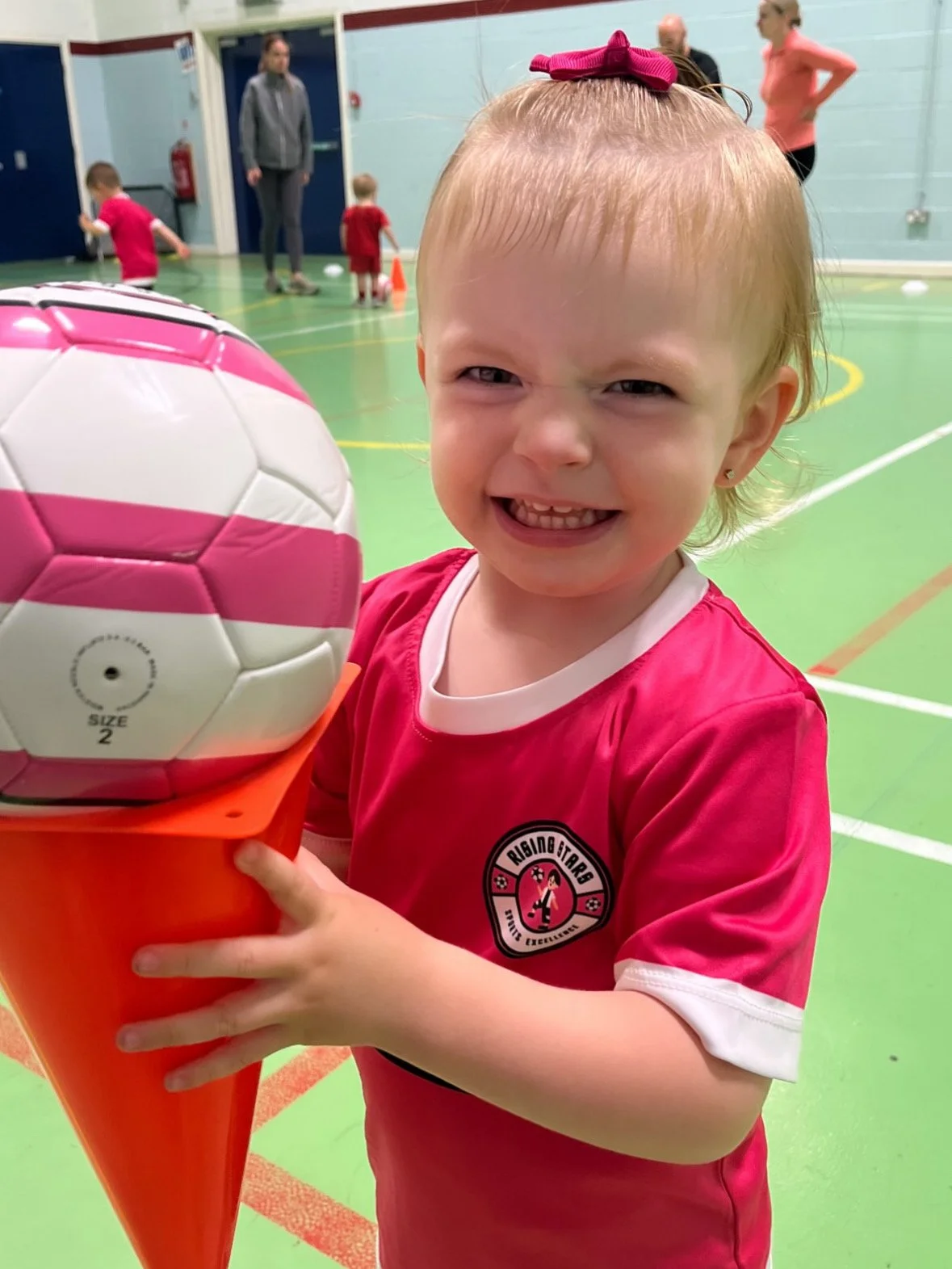 A young girl with a pink bow in her blonde hair smiling and holding a pink and white soccer ball in a gymnasium.