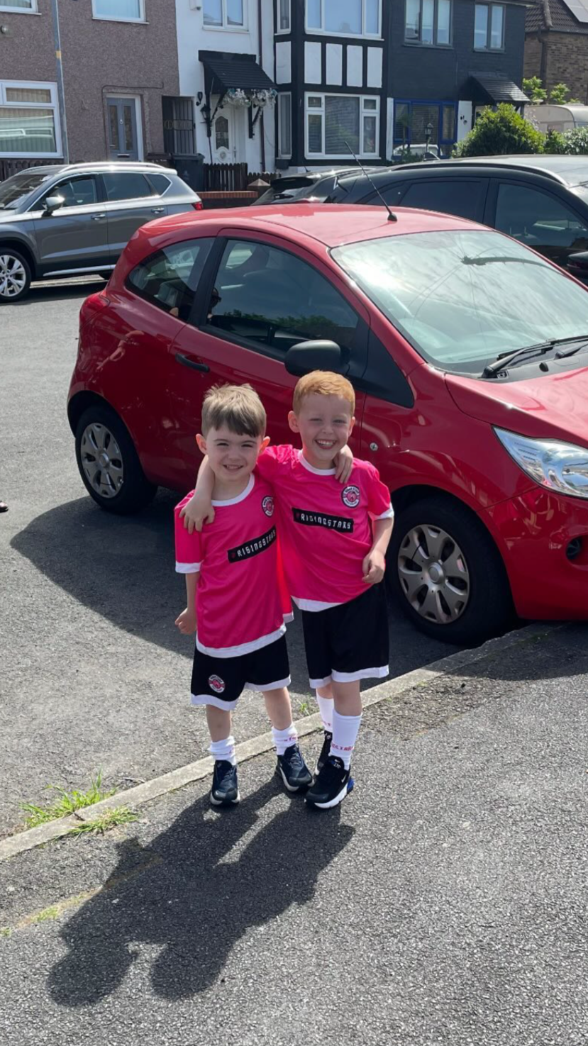 Two young boys wearing matching pink sports jerseys, black shorts, and sneakers, standing on a parking lot sidewalk with their arms around each other, smiling.