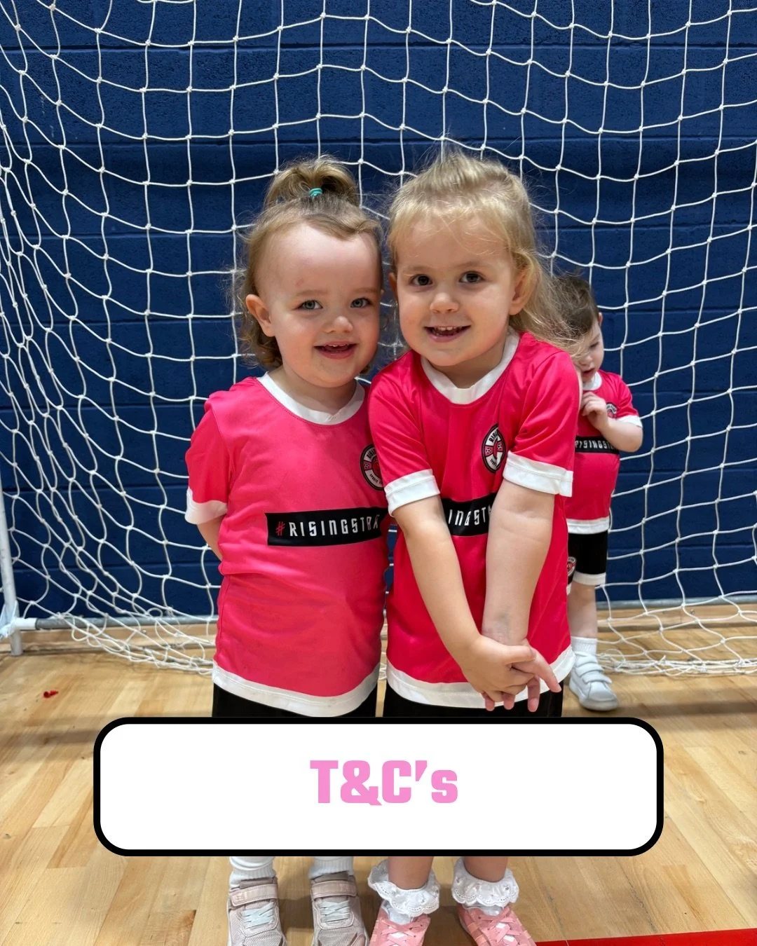 Four young girls in pink sports jerseys standing in front of a soccer goal net inside a gym, smiling.