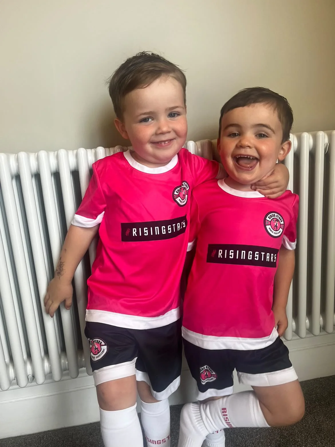 Two young boys in pink sports jerseys and black shorts, smiling and standing close together against a radiator, with one child's arm around the other's neck.