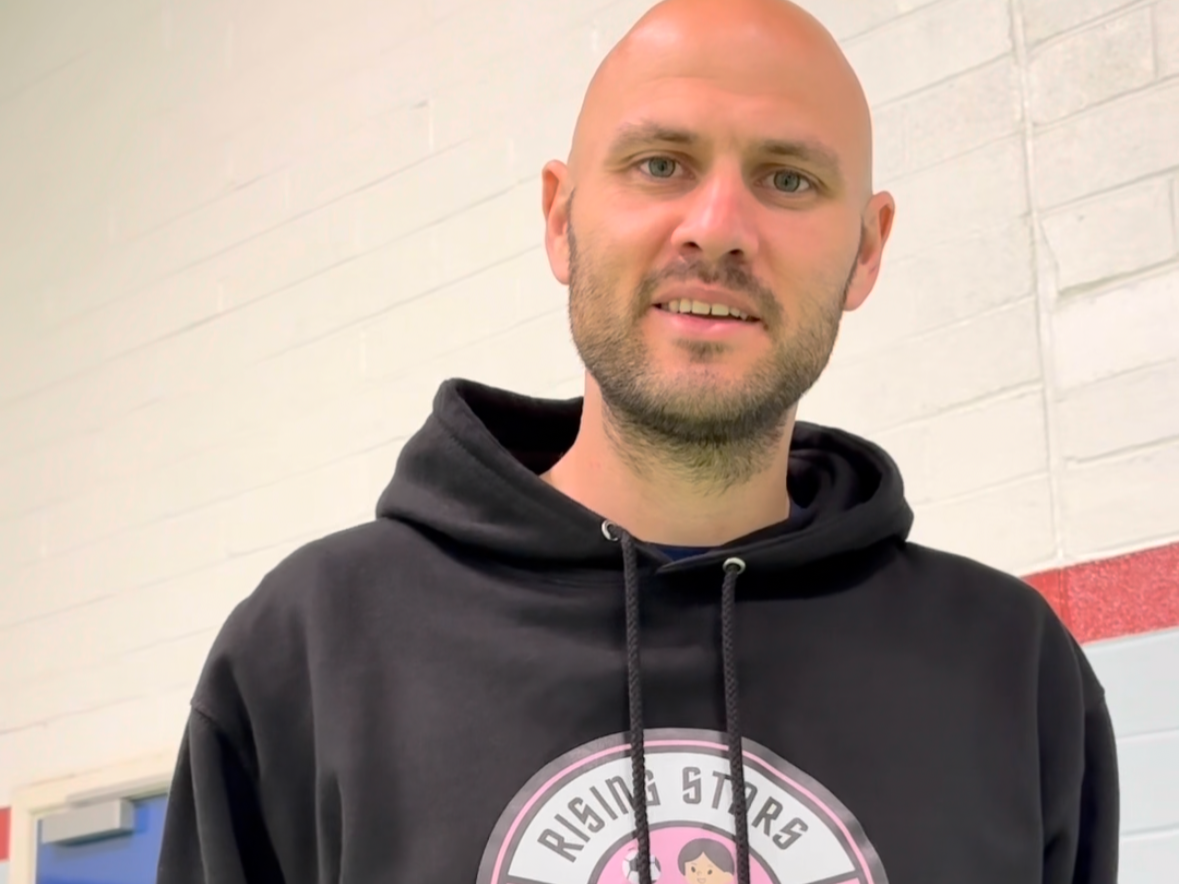 A man with a beard and shaven head standing in front of a white wall, wearing a black hoodie with a pink and white logo that says 'Rising Stars'.