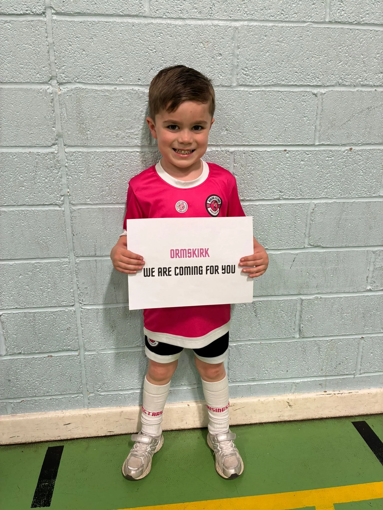 A young boy wearing a red and white sports uniform, white socks, and silver sneakers, standing indoors against a gray cinder block wall, holding a sign that reads 'ORMSKIRK' and 'WE ARE COMING FOR YOU'.