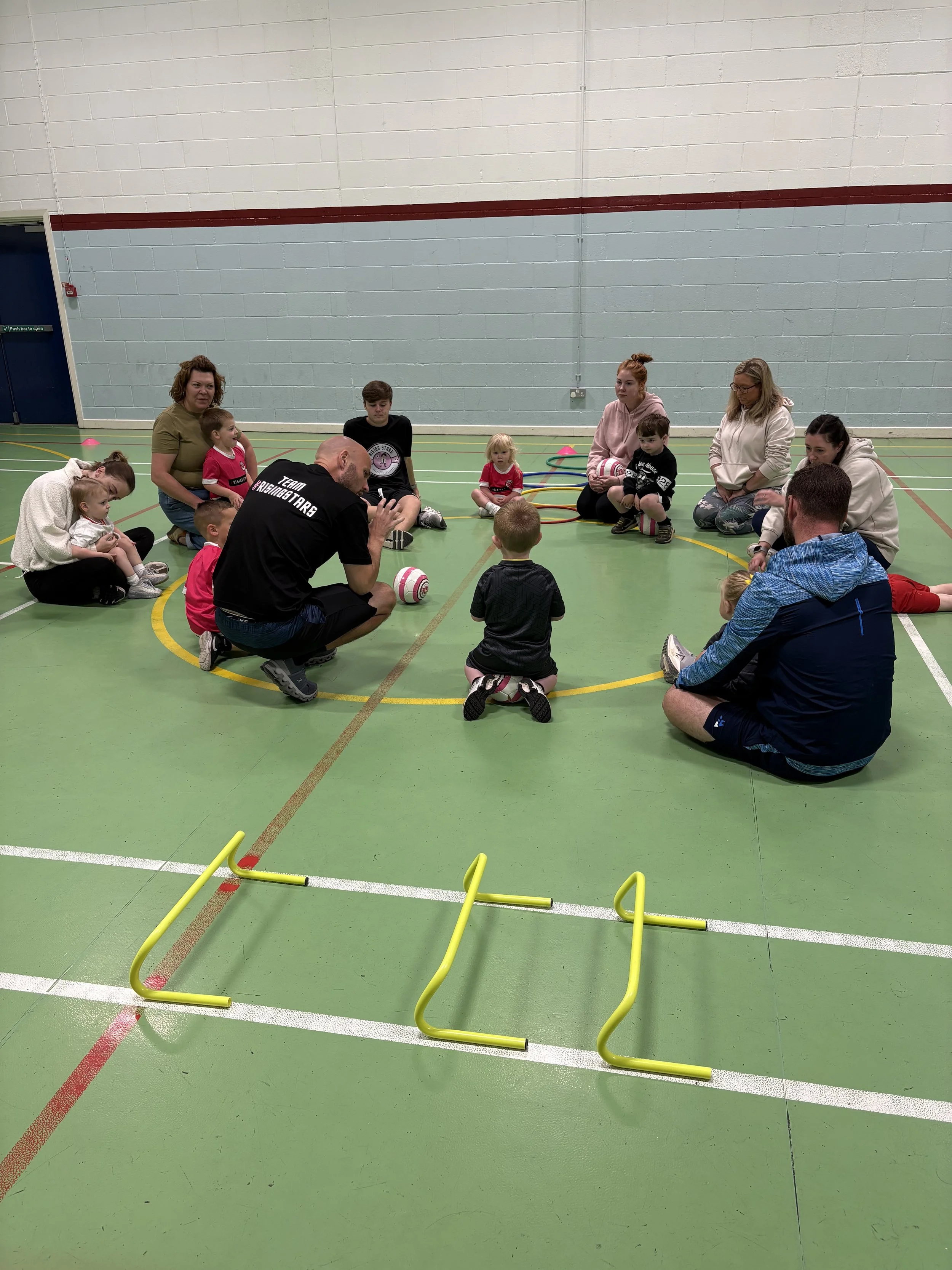 Children and adults sitting in a circle on a gymnasium floor, participating in a children’s sports or activity class, with small hurdles placed in front.