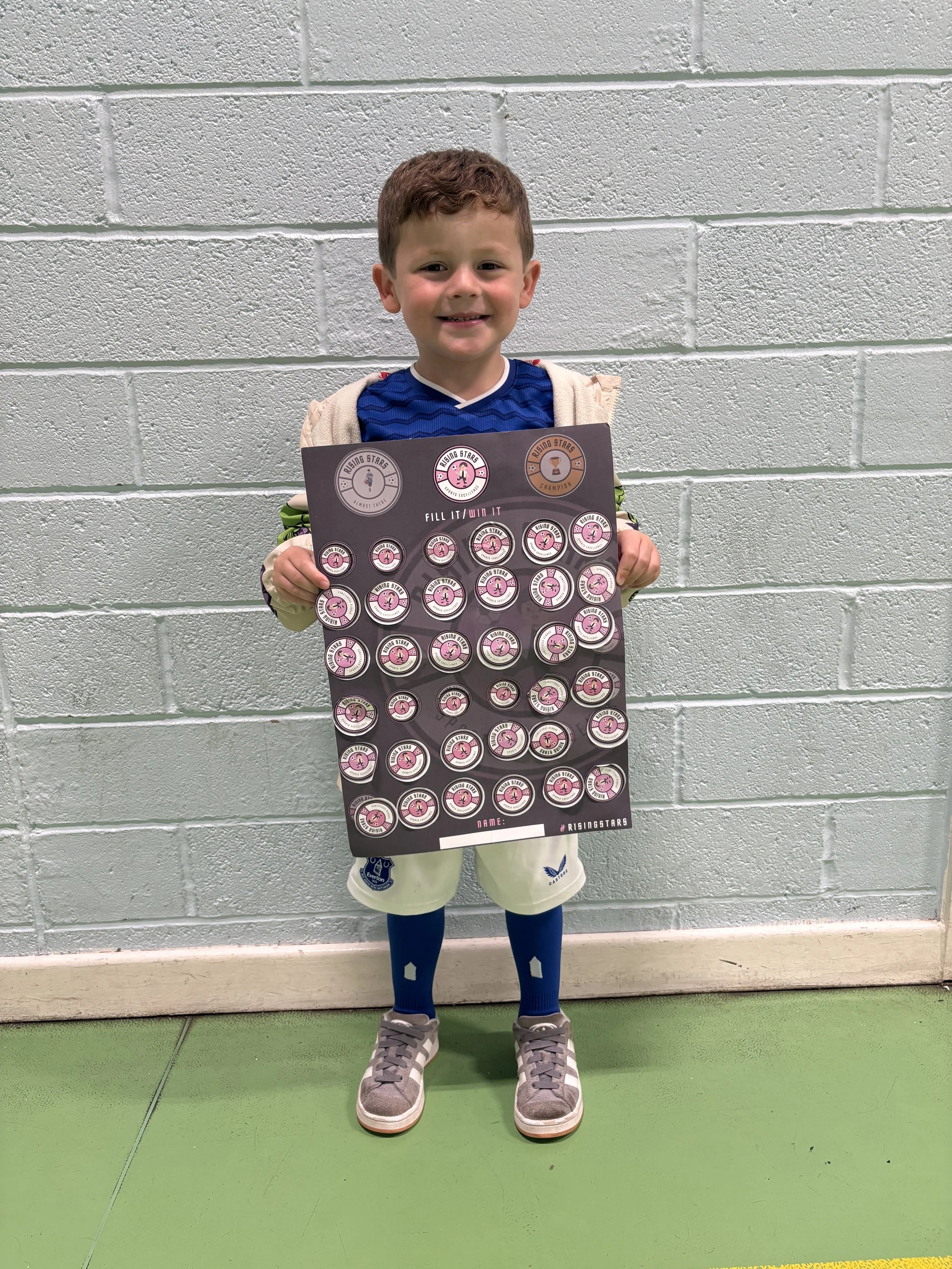 A young boy smiling and holding a sheet of pink star stickers in front of a light gray brick wall.