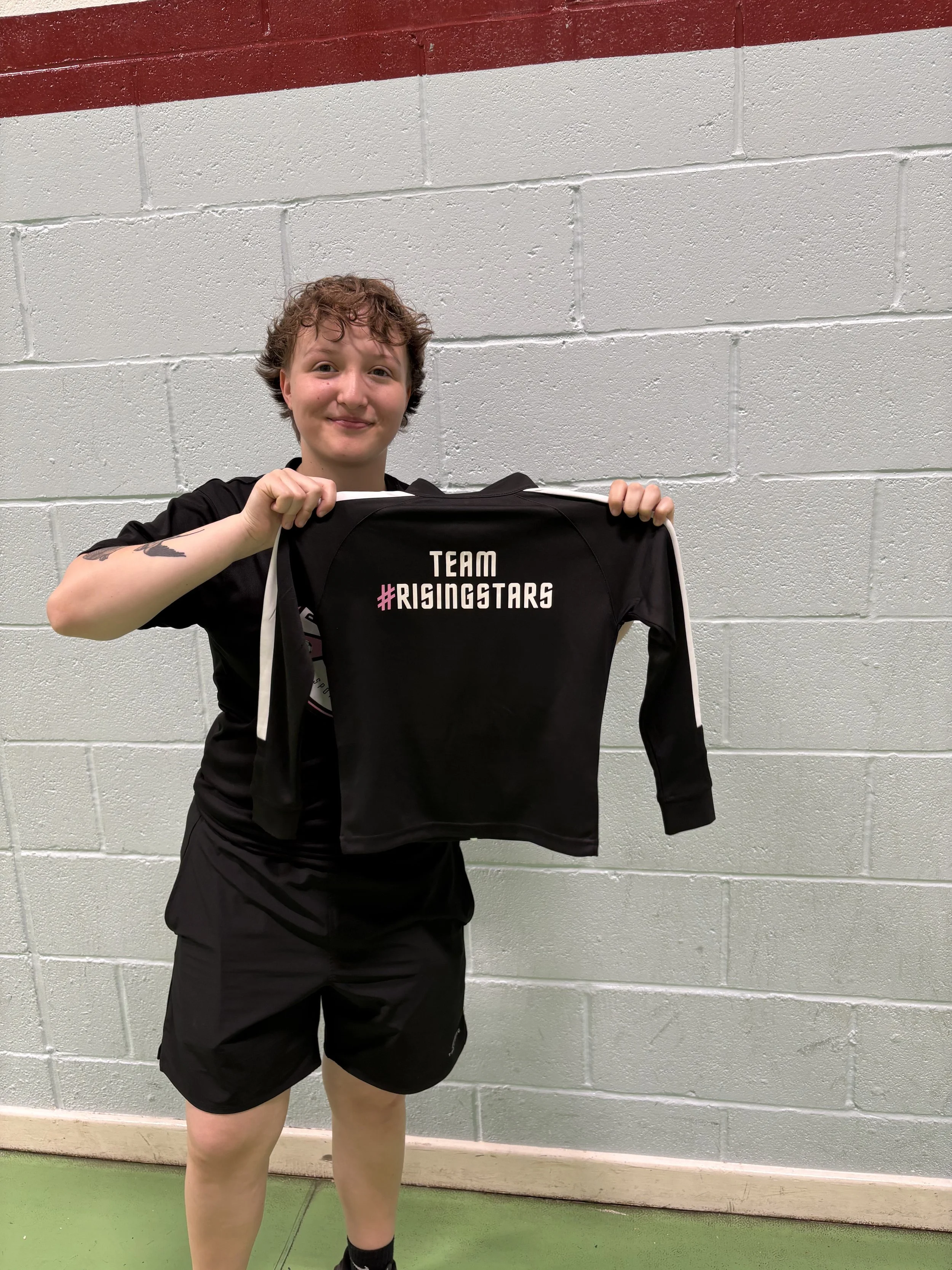 A young person with curly hair holding a black team sweatshirt that says 'TEAM #RISINGSTARS' in front of a painted brick wall.
