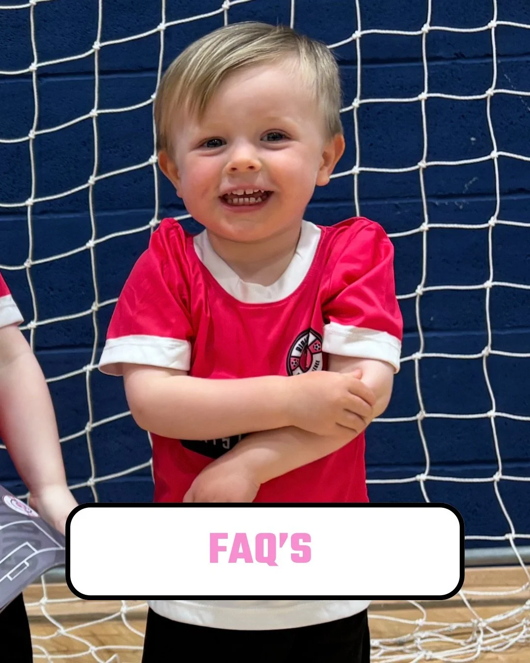 A young boy with blonde hair smiling, wearing a red soccer jersey, standing in front of a soccer goal net.