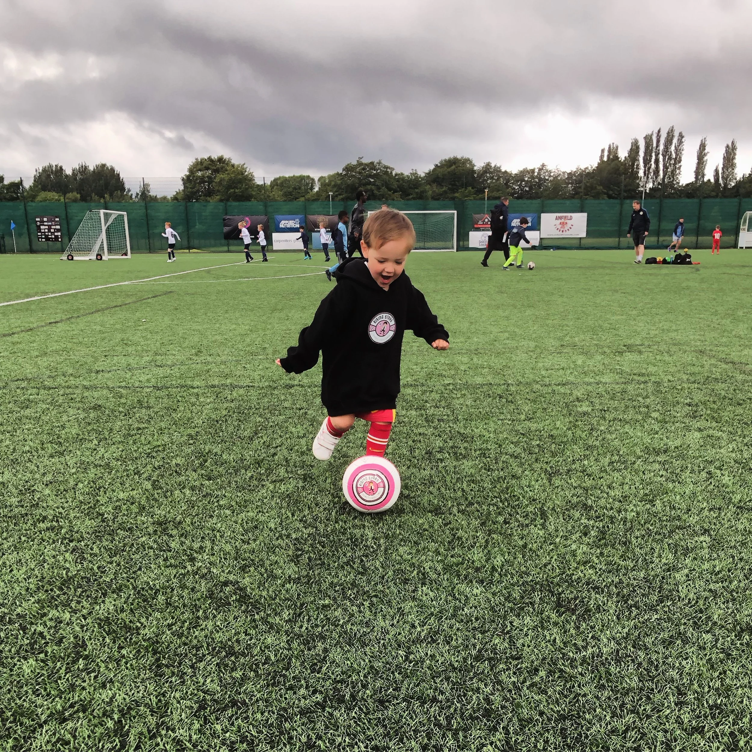 A young child playing soccer on a field, kicking a pink and white ball with other kids and adults in the background under a cloudy sky.