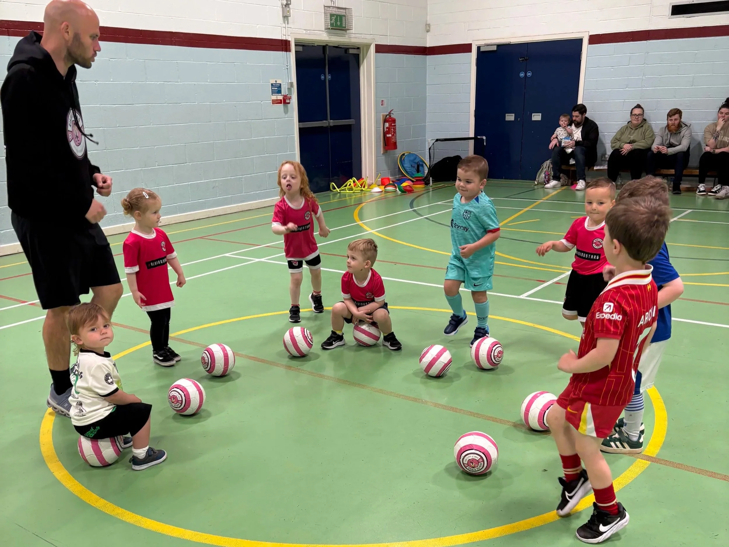 Young children participating in a soccer training session indoors, with a coach guiding them. The children are gathered around basketballs on a green indoor court, some standing and others sitting or kneeling, dressed in colorful sports uniforms.