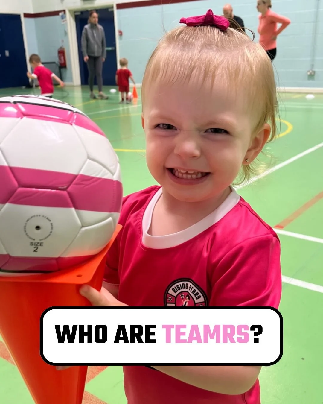 A young girl in a pink sports uniform holding a pink and white soccer ball. She has a pink bow in her hair and is smiling at the camera. In the background, there are other children and adults in a gymnasium.