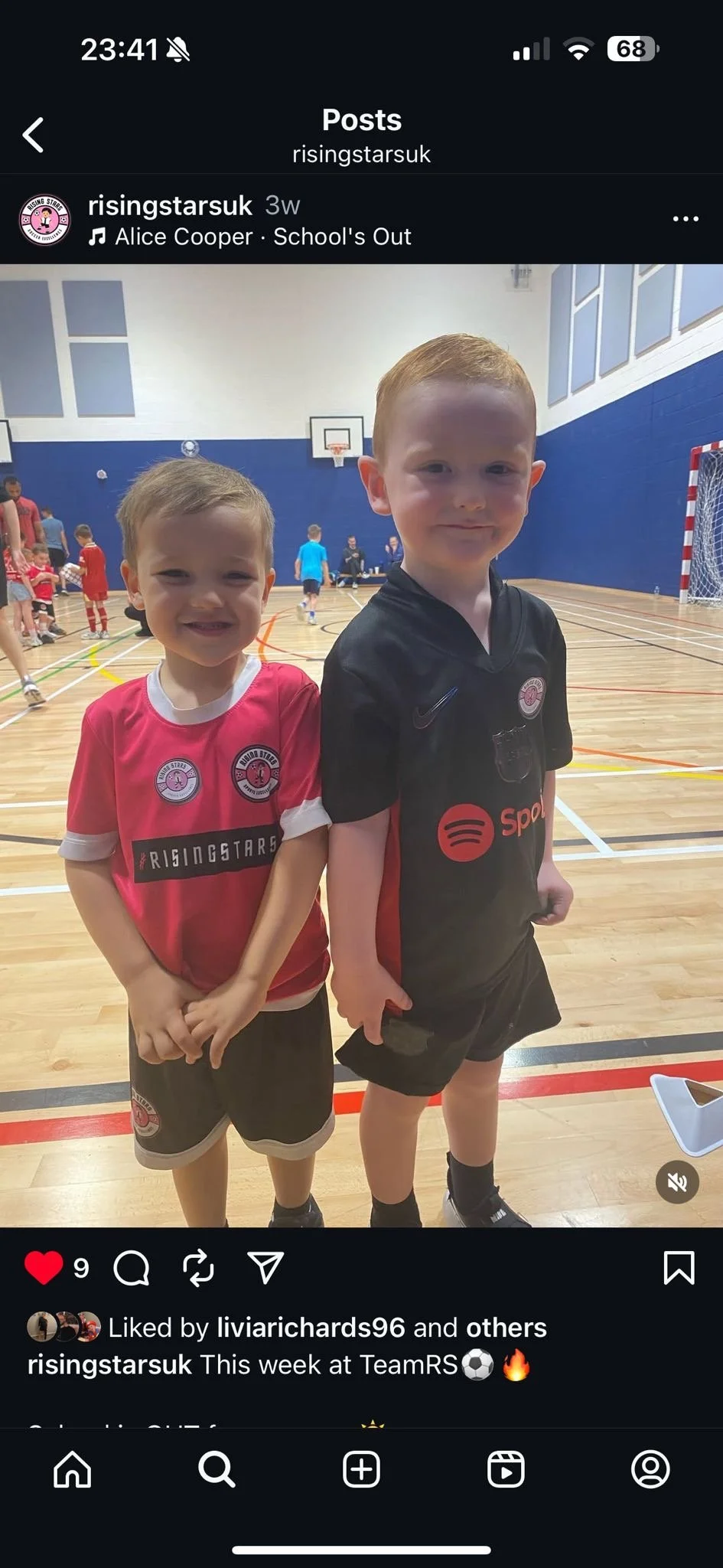 Two young boys in sports uniforms standing inside a gymnasium, smiling at the camera. The boy on the left wears a red shirt with team logos, and the boy on the right wears a black shirt with sports branding. Other children and adults are visible in the background.