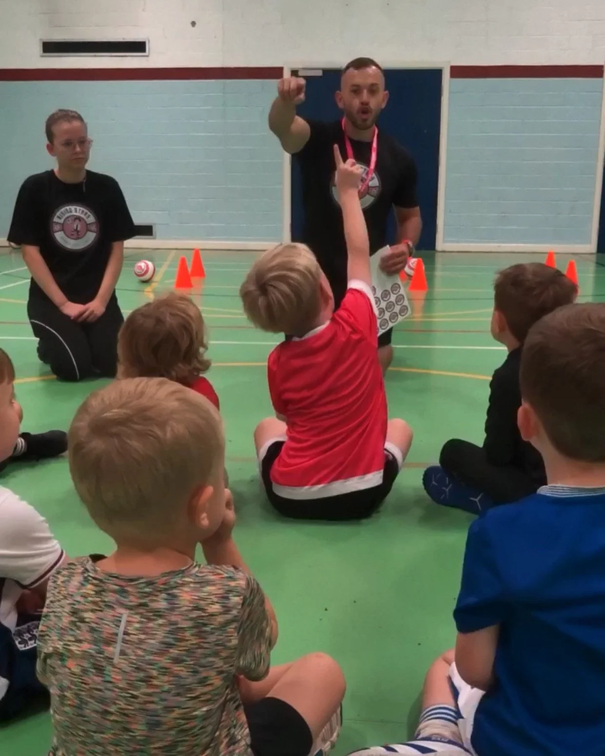 A coach in a gymnasium speaking to a group of young children seated on the floor, with some children raising their hands. There are orange cones and a rugby ball in the background.