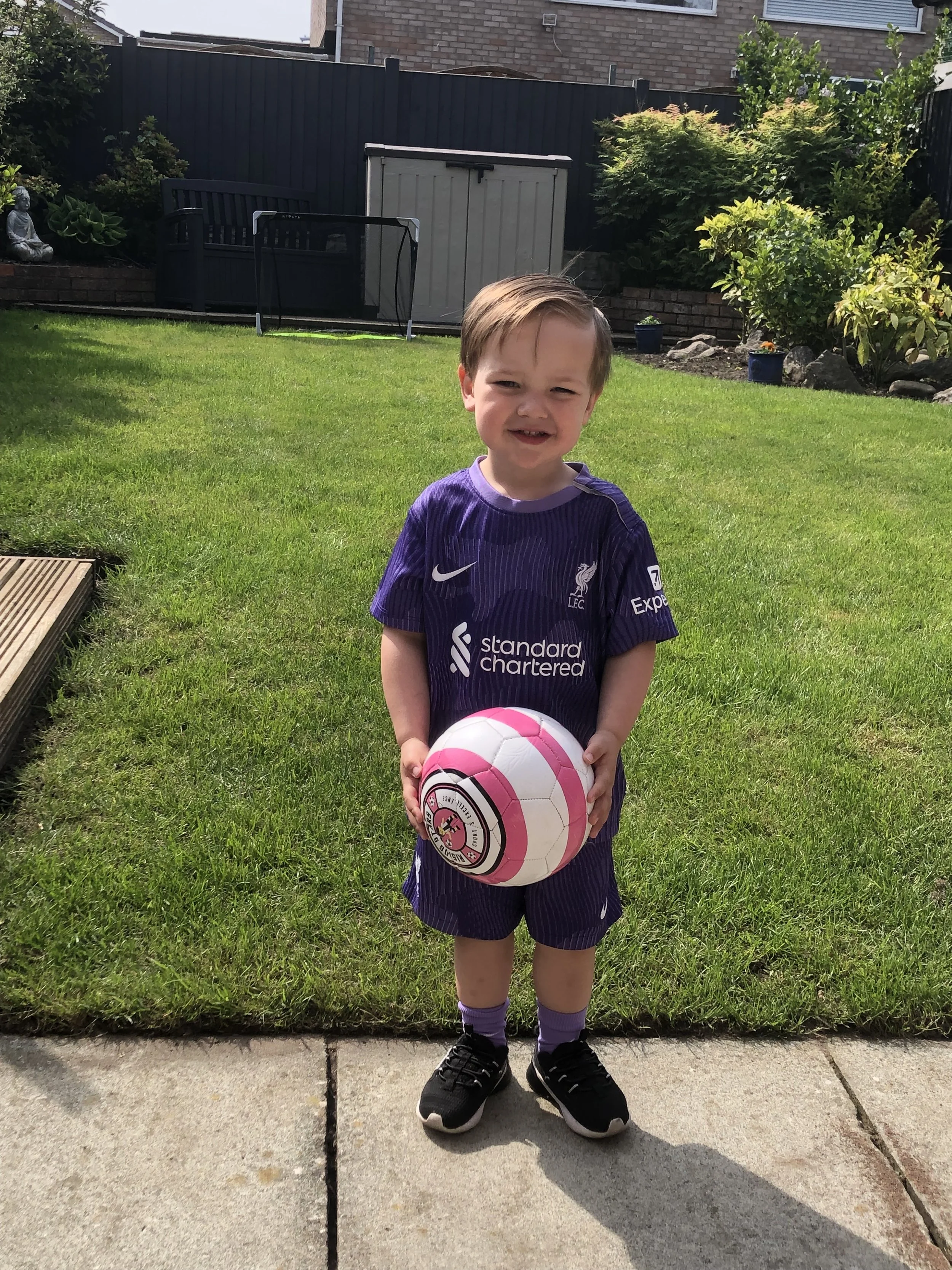 A young boy in a purple soccer jersey and shorts holding a pink and white soccer ball, standing on a sidewalk in a backyard with grass and plants.