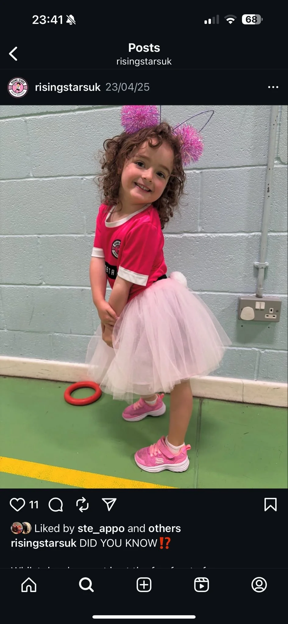 A young girl with curly hair smiling, wearing a pink T-shirt, a pink tutu, pink sneakers, and a headband with pink pom-poms and antennae, standing indoors against a light gray brick wall.