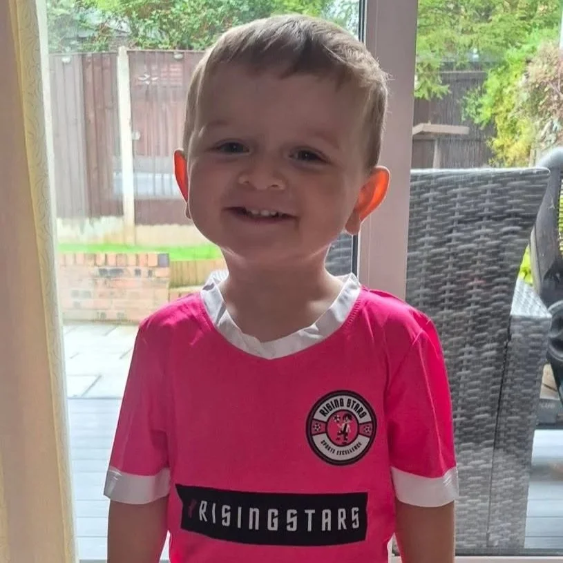 A young boy with short light brown hair, smiling and showing his teeth, standing indoors in front of a glass door, wearing a pink sports jersey with a white collar and sleeve cuffs, and there is a patio with outdoor furniture and greenery visible outside.