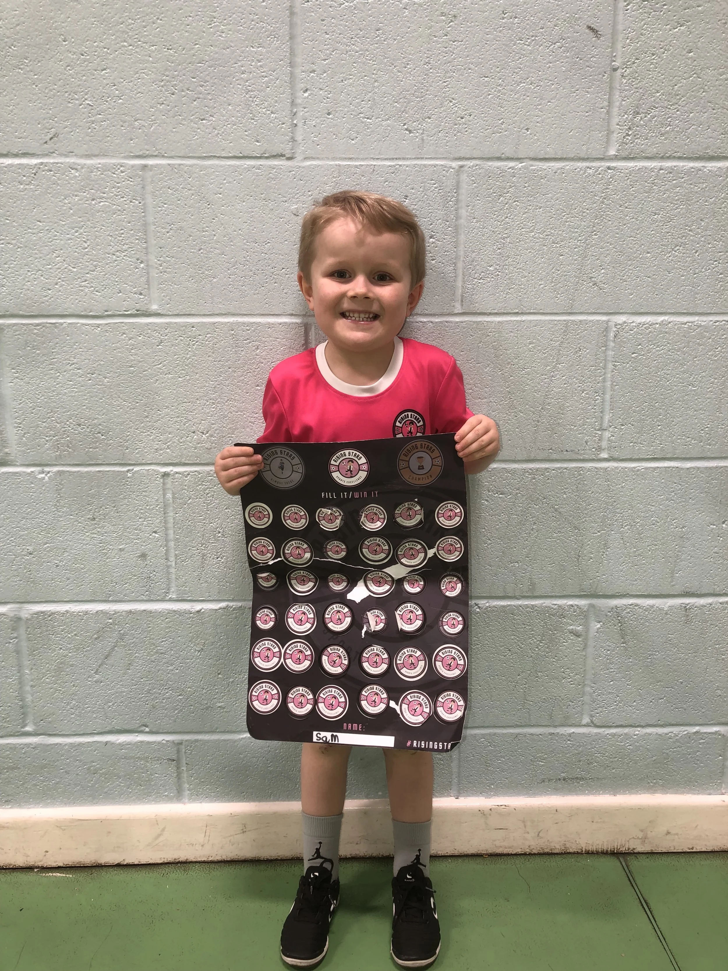A young boy standing against a light green wall, smiling, holding a soccer achievement poster, wearing a pink sports jersey, grey socks, and black sneakers.