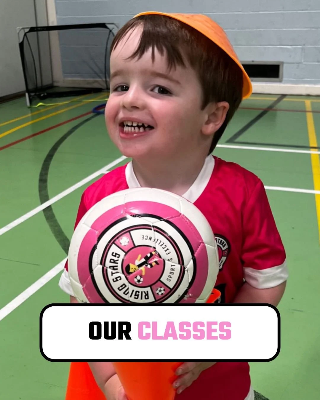 A smiling young boy with missing front teeth wearing a red soccer jersey, holding a pink and white soccer ball, inside a gymnasium or sports hall.