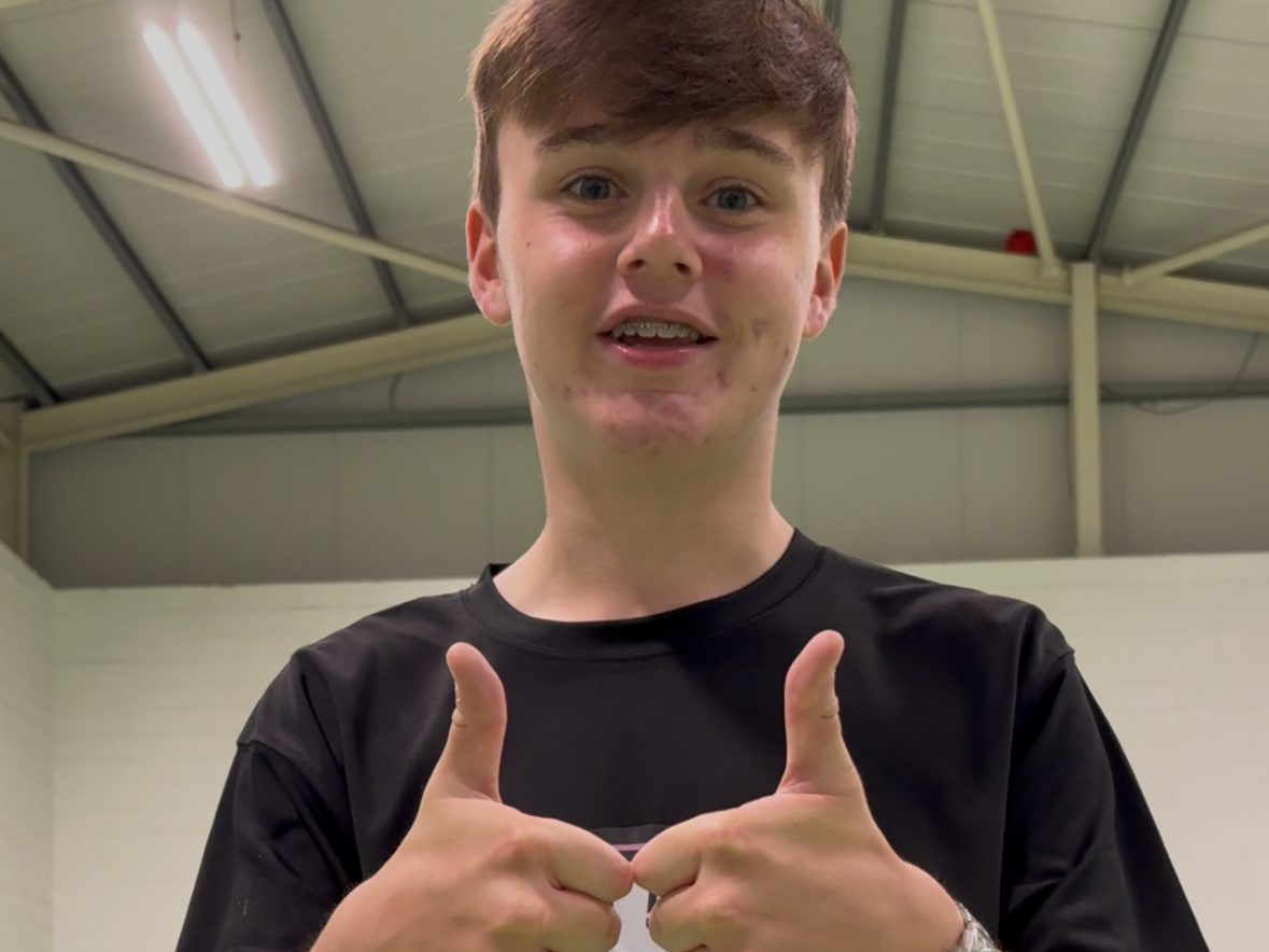 A young man with light skin and brown hair giving two thumbs up indoors with a ceiling of metal beams and white walls in the background.