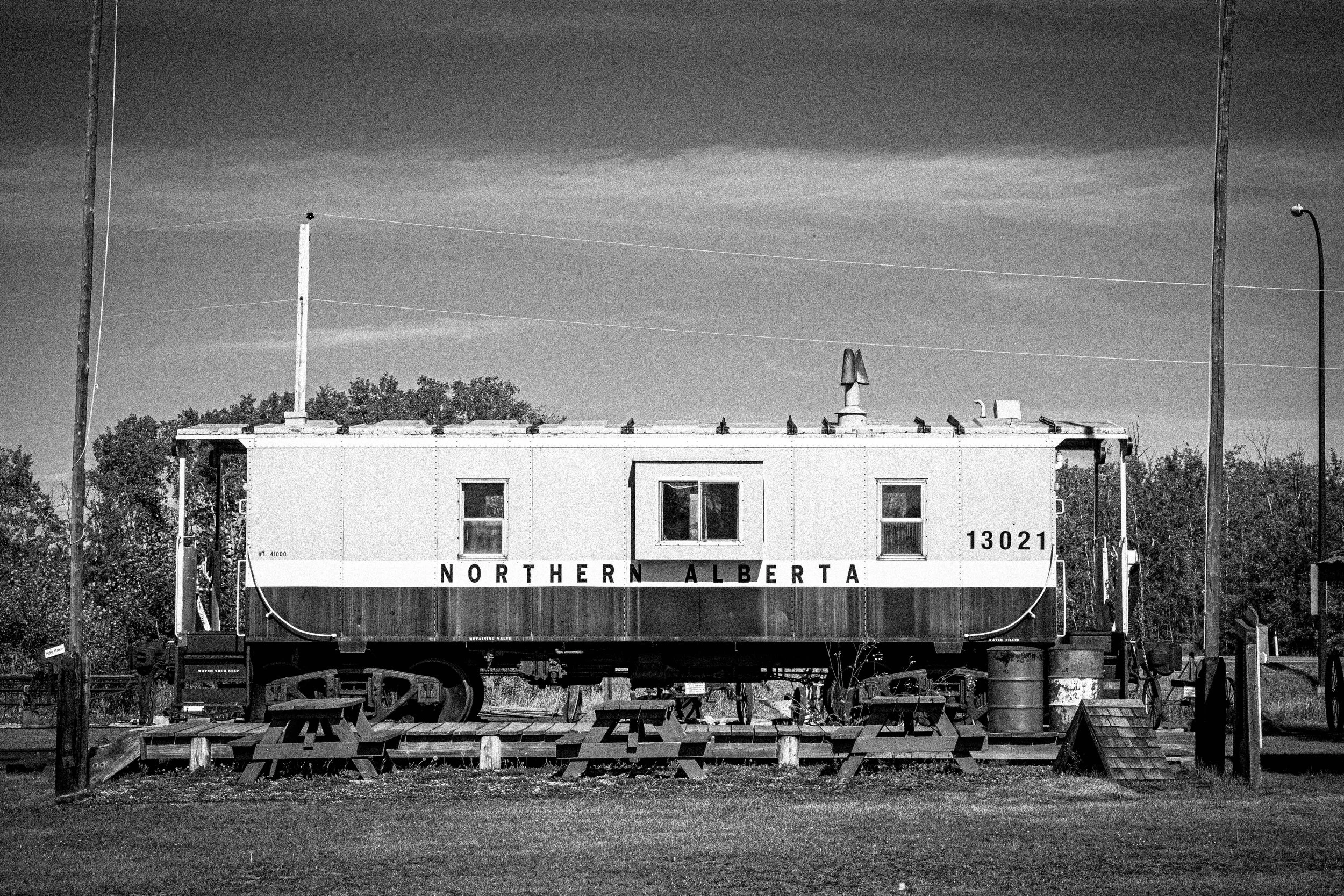 A black-and-white photo of a vintage train car with the words 'Northern Alberta' written on its side, parked on railroad tracks, with trees and utility poles in the background.