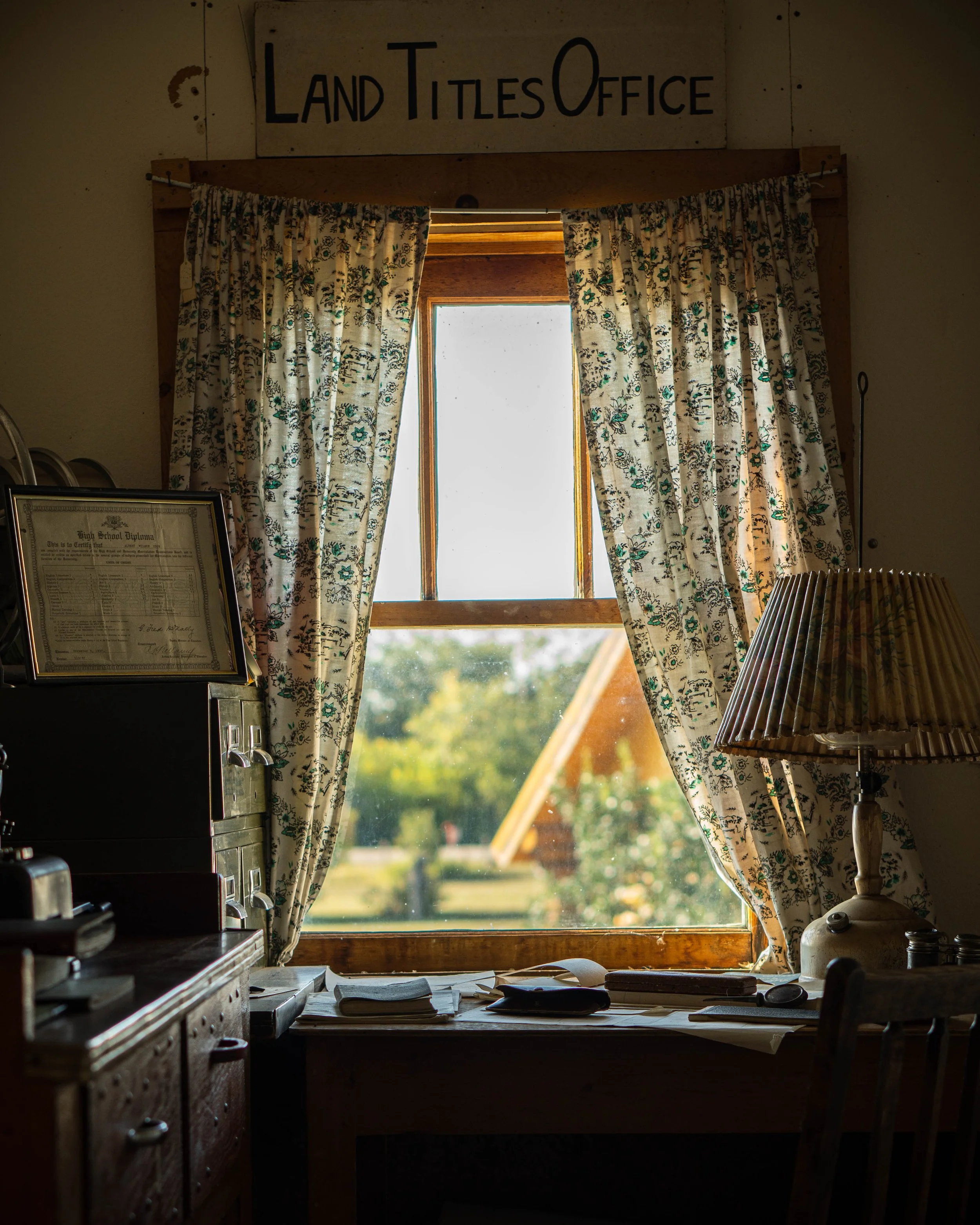 A rustic office with a sign reading 'Land Titles Office' above a window with floral curtains. The window looks out onto a rural scene with trees and a building with a sloped roof. On the desk below the window are papers, a record player, and a lamp.