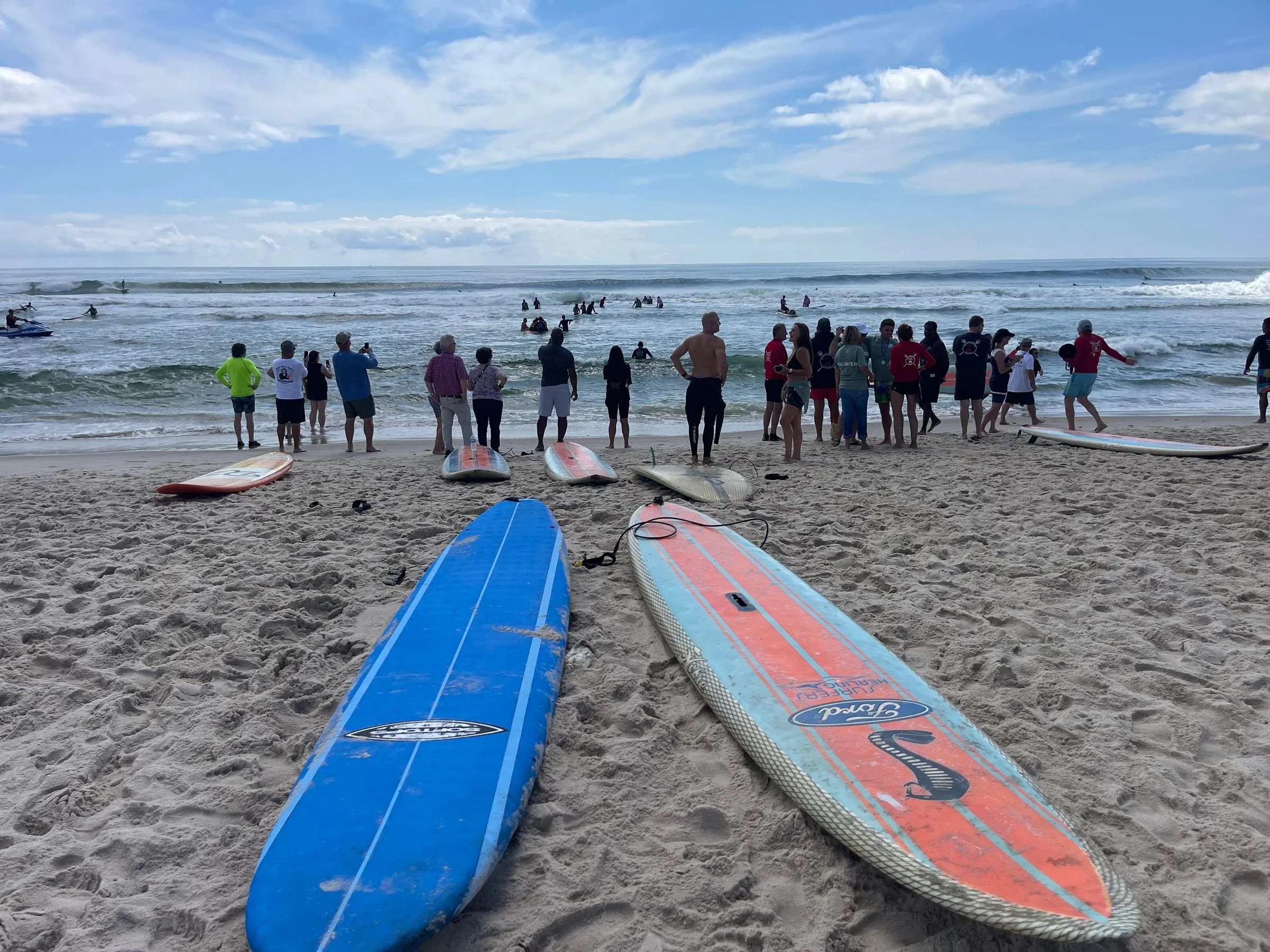 Surfboards lying on the sandy beach with people gathered near the shoreline, some preparing to surf or watching others, and the ocean with waves and people in the distance under a partly cloudy sky.