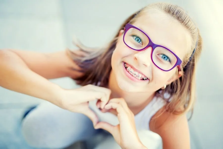 A smiling girl with blue eyes wearing purple glasses making a heart shape with her hands.