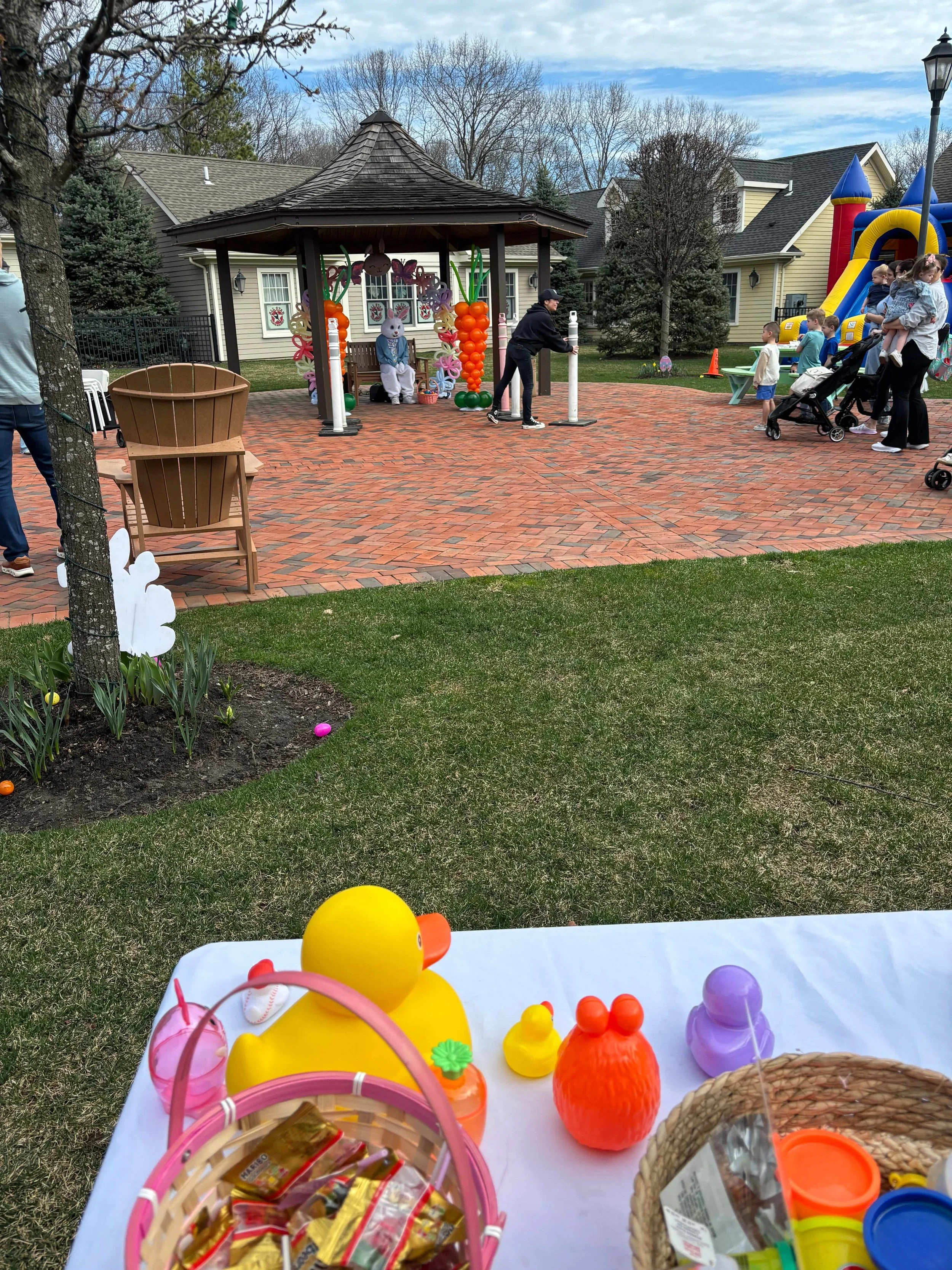 Easter celebration in a backyard with a table of plastic toys and Easter eggs in the foreground, a small decorated gazebo, a slide, and children and adults enjoying the event in the background.