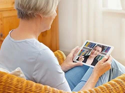 Older woman sitting in a wicker chair, looking at a video call on a tablet, smiling while talking to a woman in a library.