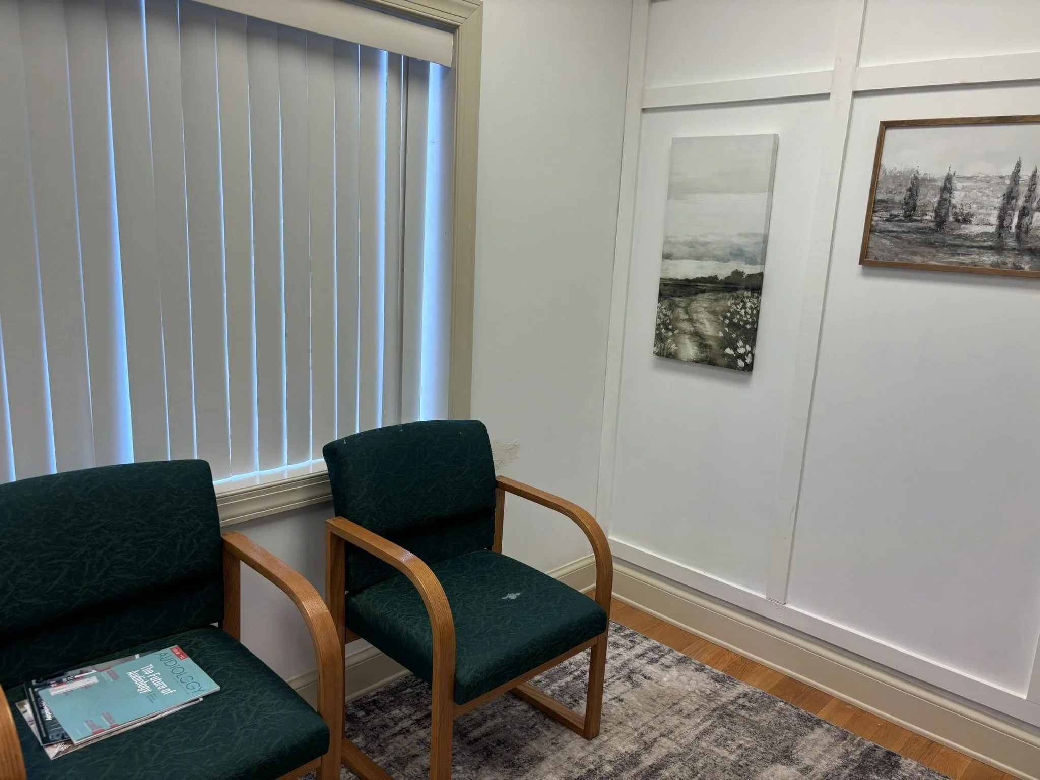 Waiting room with two green chairs with wooden arms, a magazine on one chair, a window with vertical blinds, and two landscape paintings on a white wall.