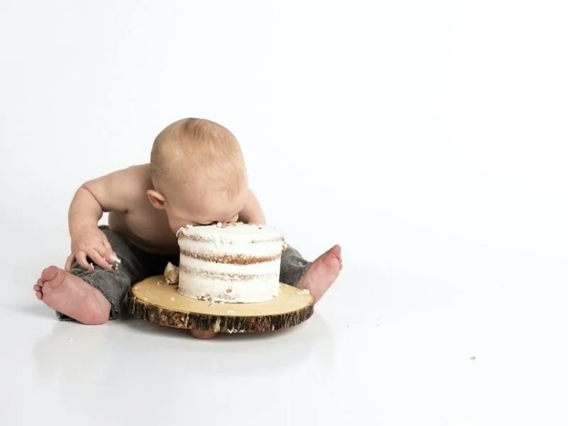 A baby with light-colored hair and no shirt sits on the floor eating a small, partially eaten cake on a wooden slab.