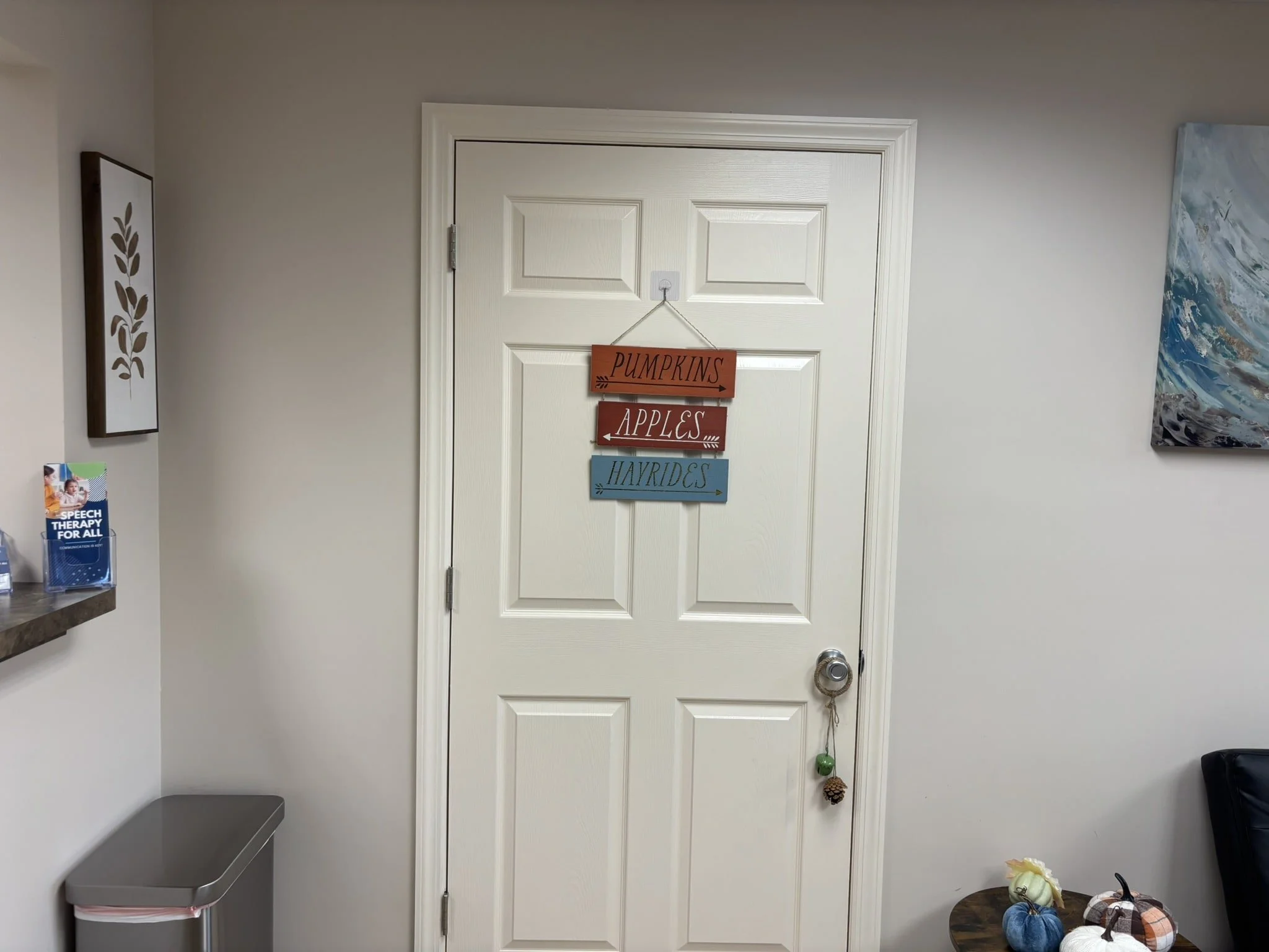 A door with a hanging sign that says "PUMPKINS APPLES HAYRIDES" in a room with light-colored walls. To the left, there is a small gray trash can and a shelf with pamphlets. To the right, there is a table with decorative pumpkins and a black chair. Th