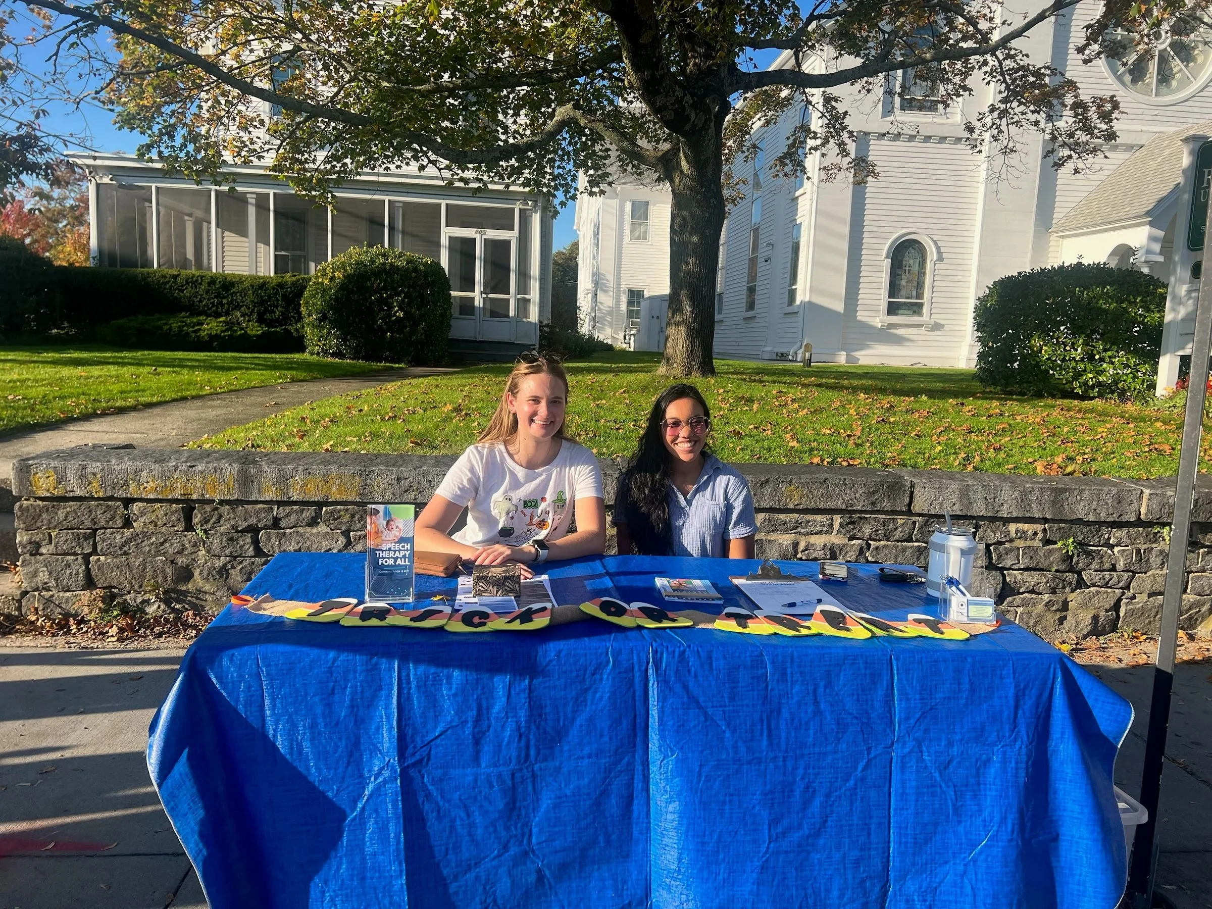 Two women sitting behind a blue tablecloth-covered table outdoors on a sunny day, with signs and materials related to speech therapy, in front of a stone wall and a white church building with trees and bushes.