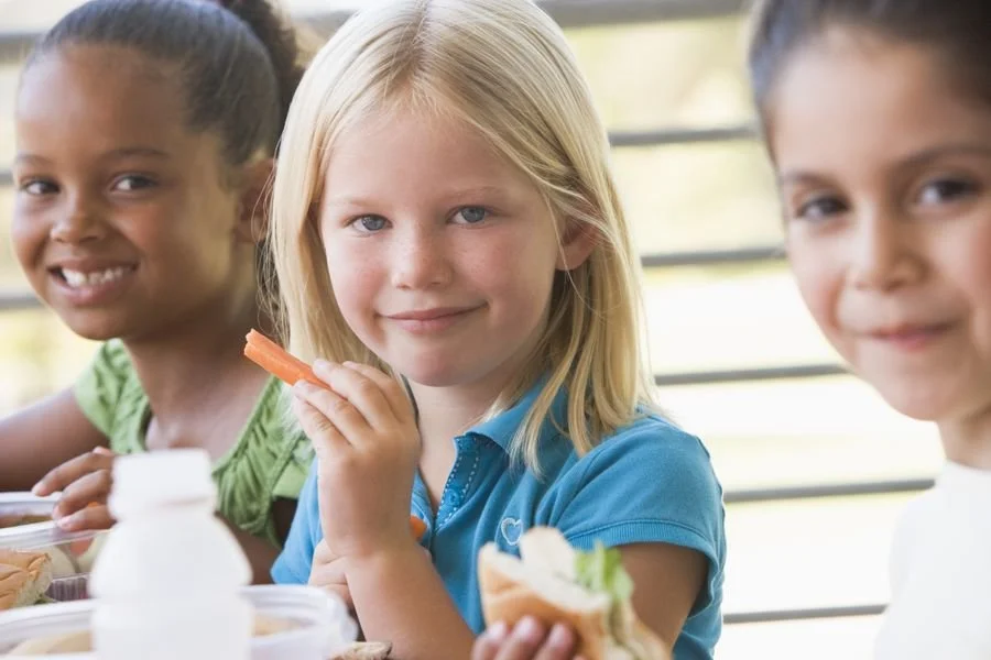 Three young girls sitting at a table, enjoying a meal outdoors, with the focus on the blonde girl in the middle holding a carrot stick.