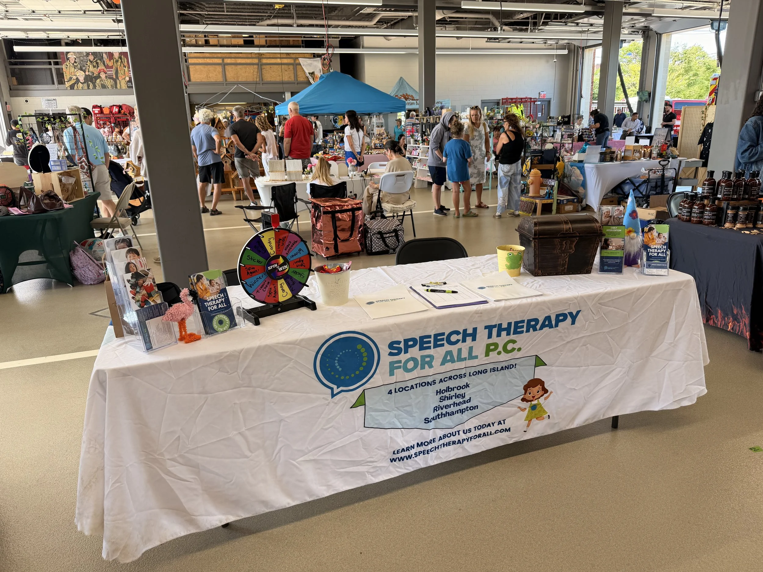 Speech therapy for all organization booth at an indoor event, with tables displaying brochures, a prize wheel, and various items for sale, and people browsing in the background.