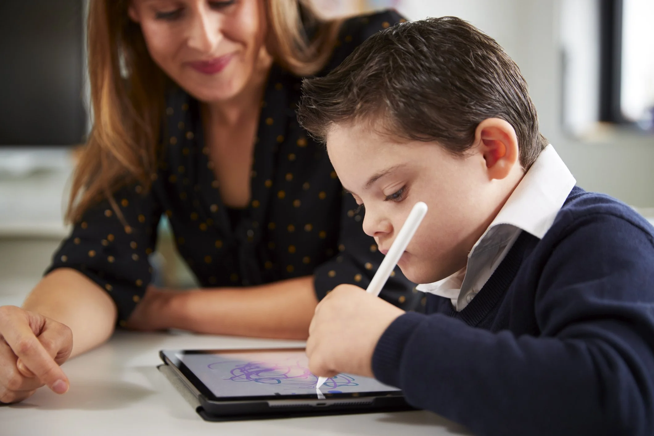 A boy in a school uniform drawing on a tablet with a stylus while a woman, possibly a teacher or parent, looks on and smiles.