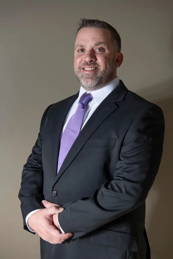 A man in a dark suit, white dress shirt, and purple tie smiling for a portrait against a plain background.
