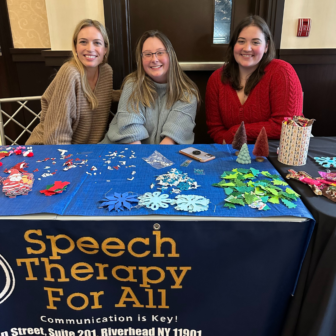 Three women sitting behind a table decorated for Christmas with various craft supplies, smiling at the camera, at a Speech Therapy event.