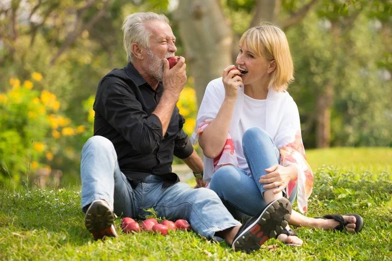 An elderly man and a young woman sitting outside on grass, enjoying apples together in a park.