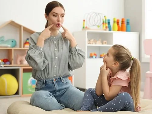 A woman with dark hair in a gray shirt and jeans making faces with her tongue out and fingers in her cheeks, sitting on a cushion, while a young girl with blonde hair in a pink shirt and blue polka dot pants sits cross-legged, smiling and looking at her, in a playroom with colorful toys and shelves in the background.