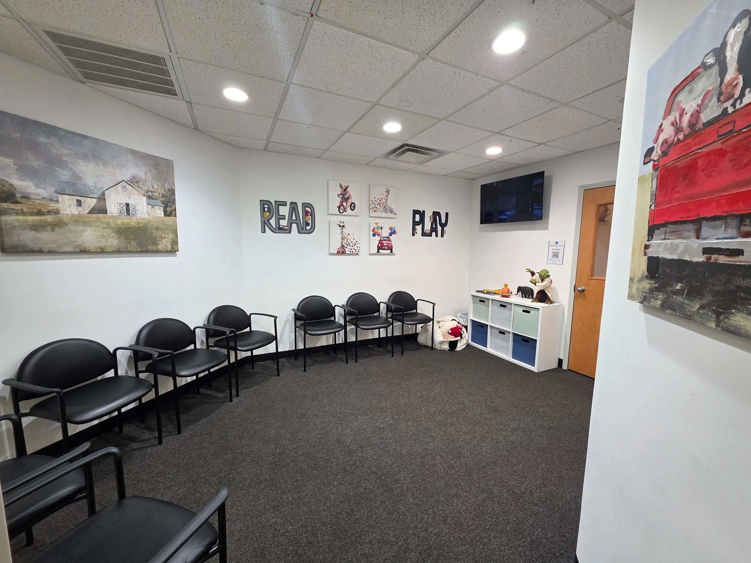 Children's waiting room with black chairs arranged along white walls, colorful wall decor including the words 'READ' and 'PLAY,' and children's artwork. There is a white storage unit with toys and a bulletin board on the wall. The room has a gray car